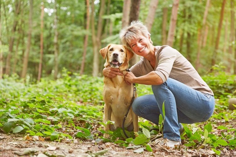 A woman with short hair kneels next to her tan colored dog while outside on a trail