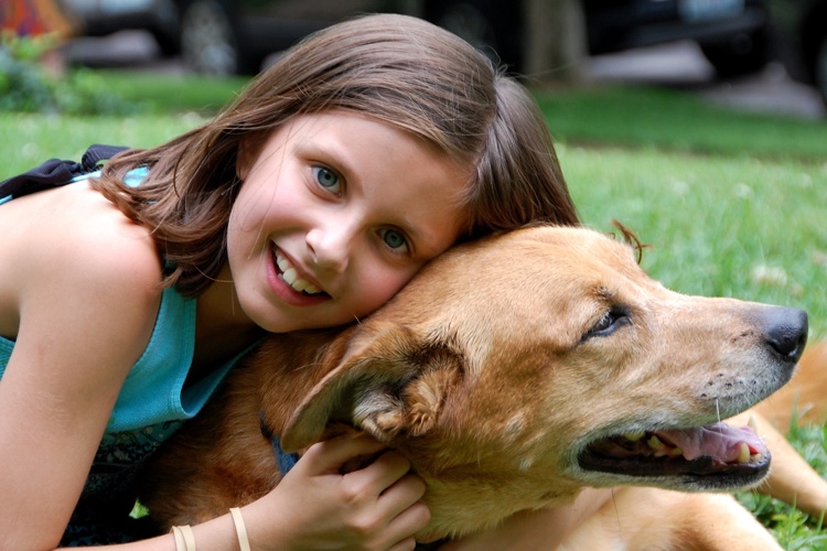 Young brunette girl sits in the grass and with her arms wrapped around a light brown colored dog